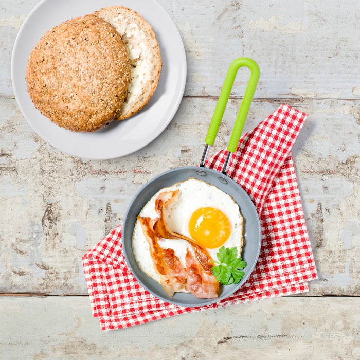 The Mini Round Egg Pan by GreenPan, made of quick-heating aluminum, holds a sunny-side-up egg and bacon on a red and white checkered cloth. A sprig of parsley accents the setting, while a white plate with a sesame seed bun sits nearby on the rustic wooden surface.