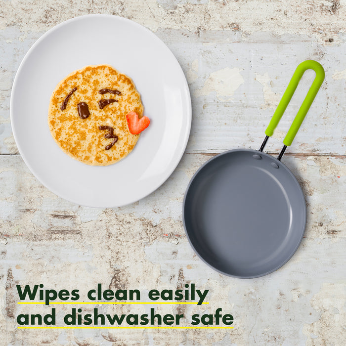 A frying pan with a green handle is placed next to a white plate holding a pancake shaped and decorated like a smiling face with a kiss mark. The text on the image reads: "Wipes clean easily and dishwasher safe."