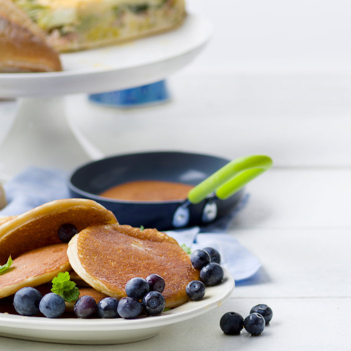 On a white table, a plate of pancakes with blueberries and mint sits beside the GreenPan Mini Round Egg Pan, featuring a green handle, quick-heating aluminum with Thermolon™ Volt coating and PFAS-free properties. In the background is a raised cake stand holding sliced cake.