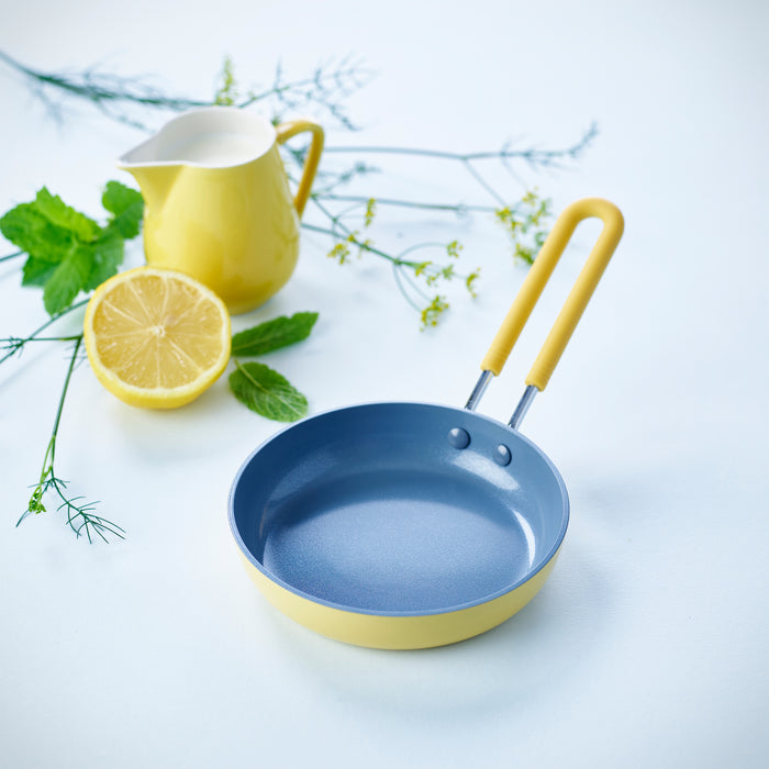 A fresh and bright scene shows a GreenPan Mini Round Egg Pan, yellow with a blue interior and PFAS-Free Thermolon™ Volt coating, on a light surface. Nearby are a matching yellow pitcher, half a lemon, and green herbs.