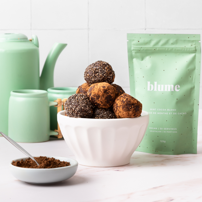 A white bowl filled with chocolate energy balls topped with chia seeds sits next to an open bag of blumes Mint Cocoa Blend, perfect for minty hot chocolate fans. A small cocoa powder bowl and pastel green teapots complement the scene on a white surface.