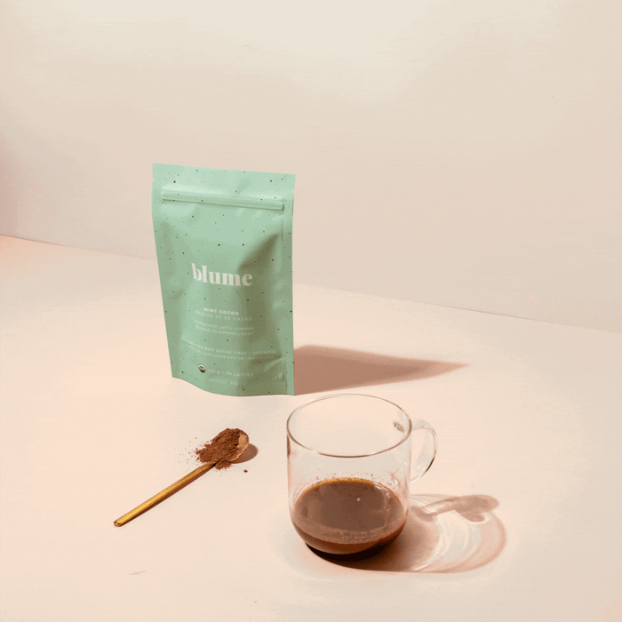 A light blue blume package stands on a surface beside a glass of minty Mint Cocoa Blend. A gold spoon with superfood powder lies nearby, set against a pale pink background.