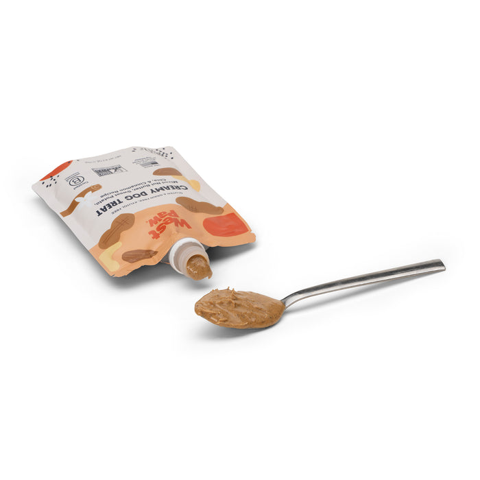 A silver spoon holds West Paws Nut Butter Sweet Potato Creamy Dog Treat beside its colorful pouch, featuring a brown and orange design with a dogs face. This delicious snack sits against a plain white backdrop.