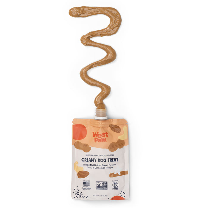 An open pouch labeled Nut Butter Sweet Potato Creamy Dog Treat reveals a wavy line of peanut butter-colored paste. The light brown packaging from West Paw is adorned with fruit and paw illustrations, highlighting its plant-based nutrition through detailed ingredient lists and certifications.