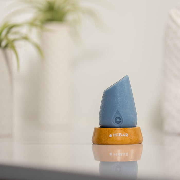 A blue, teardrop-shaped solid conditioner bar labeled HiBAR with a C sits on a wooden holder. Perfect for dry, frizzy hair, this Moisturize Conditioner contains nourishing ingredients. Blurred green plants in the background add a natural touch to the minimalistic scene.