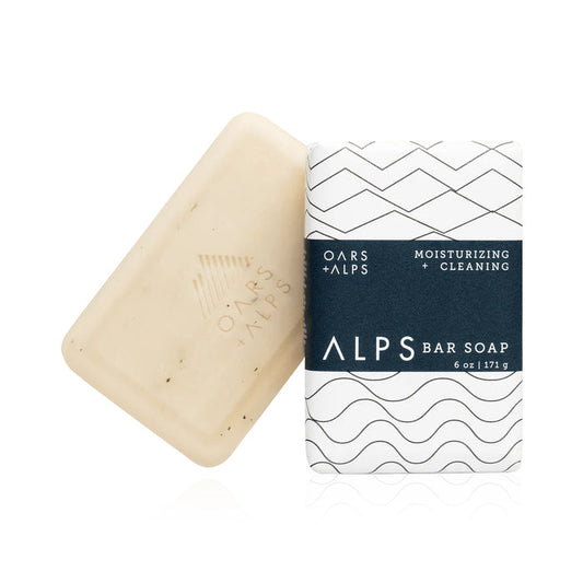 A beige Oars + Alps Moisturizing Bar Soap sits beside its white ALPS Bar Soap packaging (6 oz | 171 g), featuring a navy blue and black geometric design.