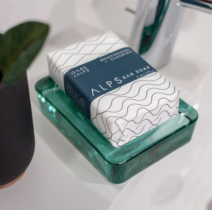 A wrapped Oars + Alps Moisturizing Bar Soap sits on a green glass soap dish beside a potted plant and chrome faucet atop a white sink.