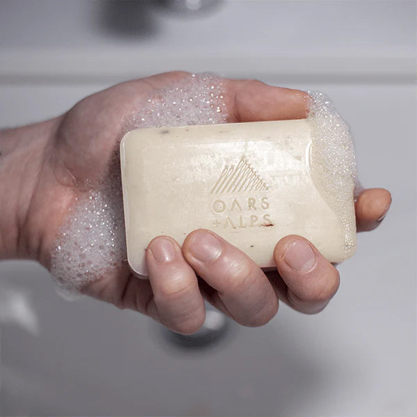 A hand holds a lathered Oars + Alps Moisturizing Bar Soap, enriched with Alpine Caribou Moss™, bubbles forming around the engraved logo, above a white sink.