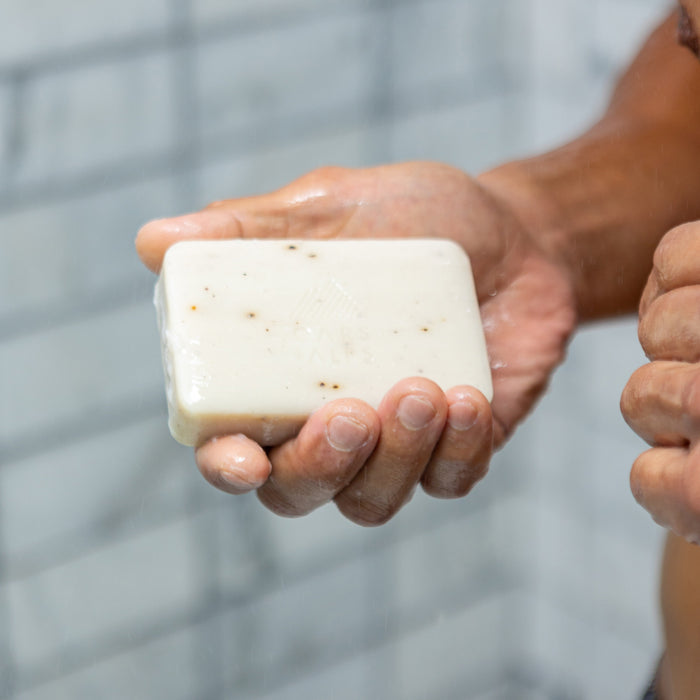 A person holding an Oars + Alps Moisturizing Bar Soap in their hand while in the shower, with a tiled wall in the background.