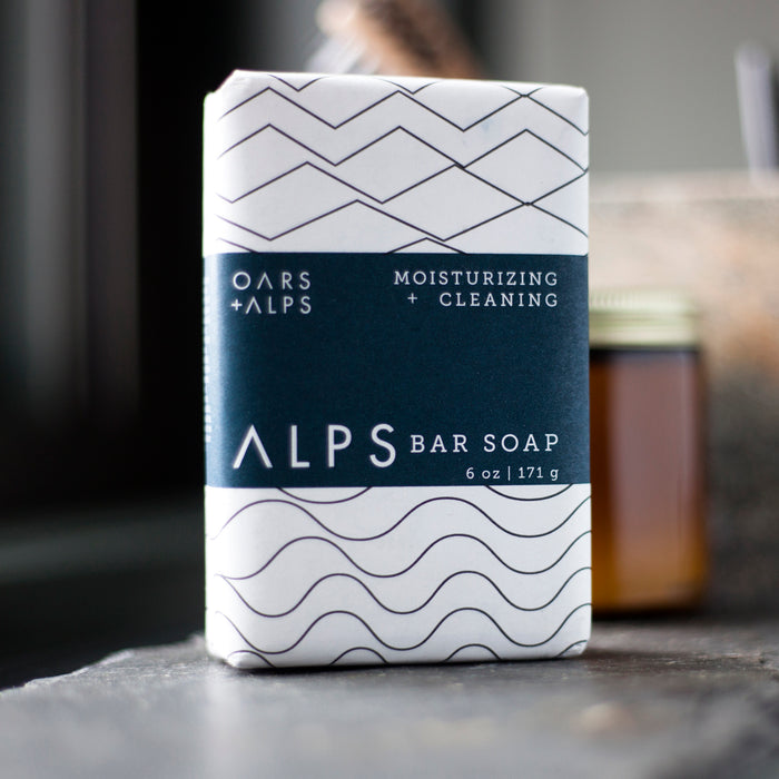 A 6 oz (171 g) Oars + Alps Moisturizing Bar Soap with nourishing Shea Butter, in white and navy wavy packaging, rests on a dark surface with a blurred jar behind it.