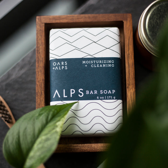 A bar of Oars + Alps Moisturizing Bar Soap with Alpine Caribou Moss™ and Shea Butter, wrapped in patterned white and navy paper, rests in a wooden tray with a green plant leaf partially covering the foreground.