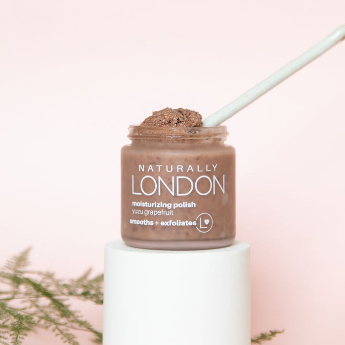 A jar of Naturally Londons Moisturizing Foot Polish with Calendula, featuring adzuki beans as a natural exfoliator, rests on a white pedestal holding a spoon. The label reads yuzu grapefruit, smooths + exfoliates. A soft pink background and green fern in the foreground complete the scene.