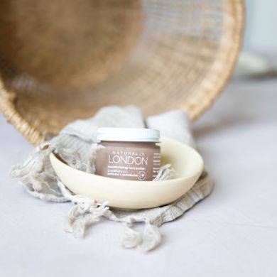 A small jar labeled Moisturizing Foot Polish with Calendula by Naturally London rests in a cream-colored bowl on fringed fabric, gracefully positioned before an upturned wicker basket, enhanced by adzuki beans natural exfoliating power.
