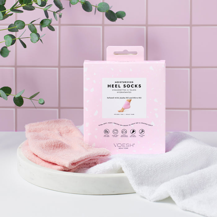 A pink box of VOESH New York Moisturizing Heel Socks infused with jojoba oil is showcased on a white circular tray with pink and white socks, set against a pink tiled wall and greenery hanging above.