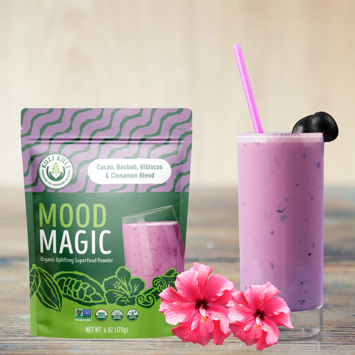 A pouch of Mood Magic Organic Superfood Blend from Kuli Kuli Foods is next to a purple smoothie with a pink straw and cherry garnish. The package highlights its cacao, baobab, hibiscus, and cinnamon blend, complemented by vibrant hibiscus flowers.
