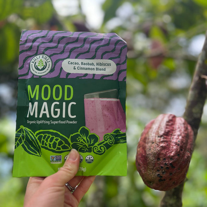 A hand holds a packet of Kuli Kuli Foods Mood Magic Organic Superfood Blend against a cacao pod, showcasing its green and purple design that highlights a unique mix of cacao, baobab, hibiscus, and cinnamon.