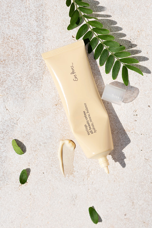 A beige tube of Ere Perez Moringa All-Beauty Crème with an open cap rests on a light textured surface, with a small amount squeezed out and green leaves scattered around, enhancing the serene skincare scene.