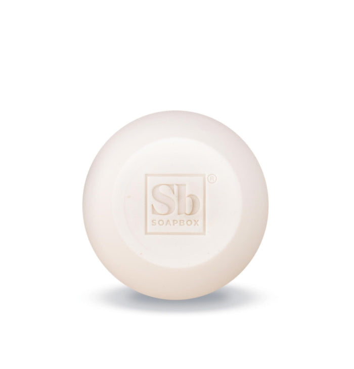 A round, white Moroccan Argan Oil Shampoo Bar by Soapbox is displayed on a white background, featuring an embossed Sb logo. This luxurious shampoo bar enriches with argan oil and supports reducing plastic waste by cutting out bottles.