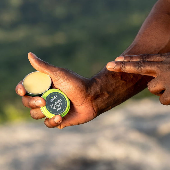 In their left hand, a person holds an open tin of Murphys Naturals Mosquito Repellent Balm, displaying the smooth balm inside. Their right hand points to highlight its DEET-free formula with natural oils, set against a softly blurred natural landscape.