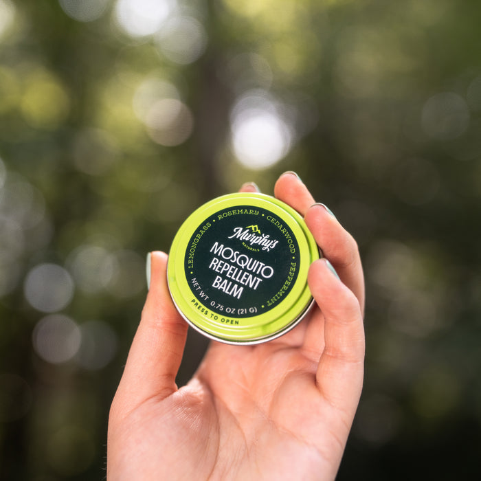 A hand holds Murphys Naturals Mosquito Repellent Balm in a small round container, featuring green with black and white text. This DEET-free balm, made with natural repellent oils, stands out against a blurred natural background as an organic mosquito deterrent.
