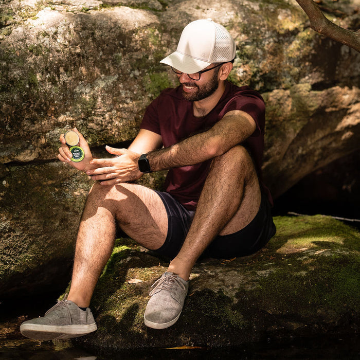 A man in a maroon shirt, shorts, and white cap sits smiling on a mossy rock by water, holding a small beverage bottle. He enjoys the shade under trees with confidence in his DEET-free Murphys Naturals Mosquito Repellent Balm. Sunglasses and a smartwatch enhance his relaxed look.
