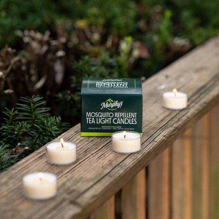 A green box of Murphys Naturals Mosquito Repellent Tea Light Candles, featuring natural soy and Citronella Oil, sits on a wooden railing with four glowing tea lights. In the background, softly blurred greenery completes the serene setting.
