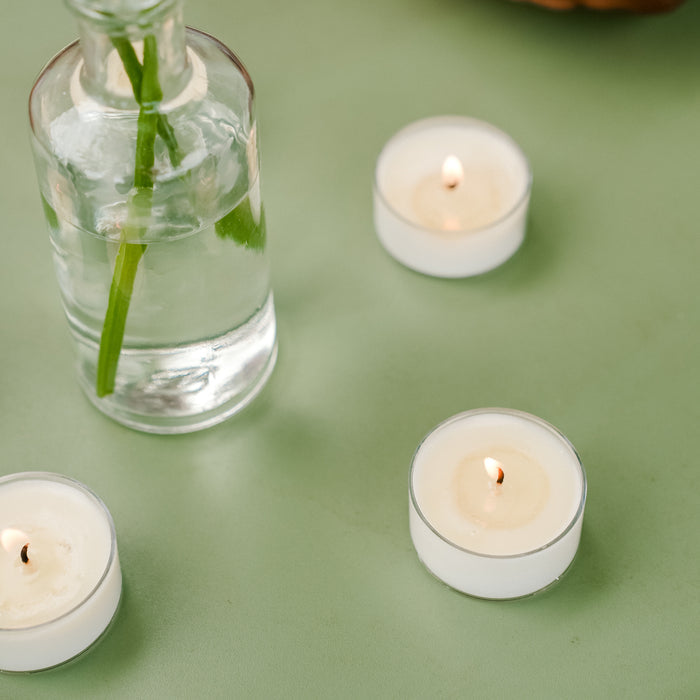 A glass vase with green stems in water sits on a green surface beside three lit Murphys Naturals Mosquito Repellent Tea Light Candles. The white, round candles, made from natural soy and citronella oil, cast a warm glow while doubling as effective mosquito repellents.