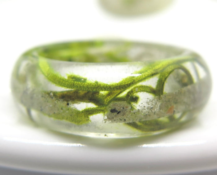 Green moss ring in clear resin with embedded ash and moss strands resting on a white surface.