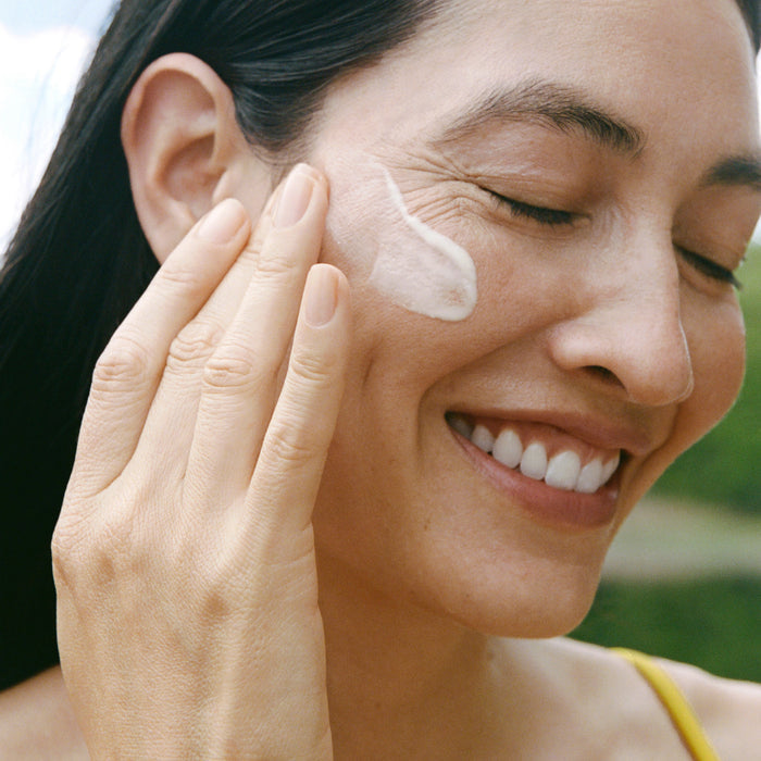 A woman with long dark hair, outdoors in a yellow top, smiles as she applies Ursa Major’s Mountain Glow Serum to her cheek with her fingers.
