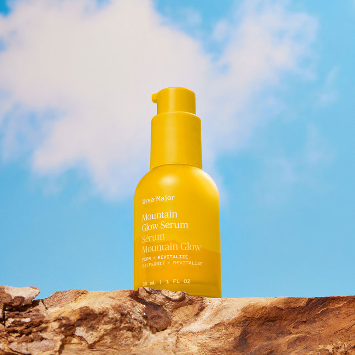 A vibrant yellow bottle of Ursa Major Mountain Glow Serum rests on a rocky surface, set against a backdrop of blue sky and gentle clouds.