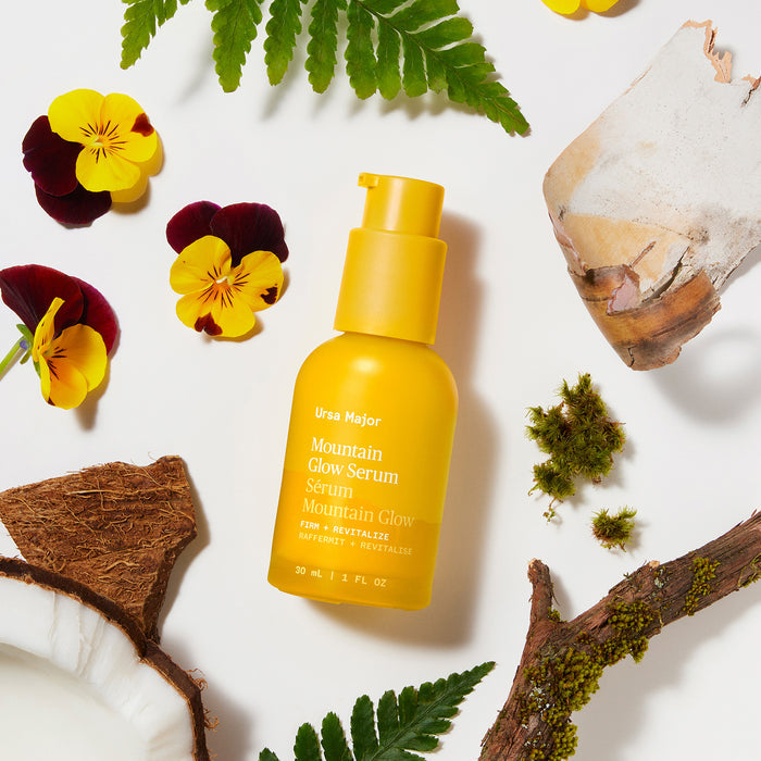 A yellow bottle of Ursa Major Mountain Glow Serum sits amid flowers, moss, a wood piece, a stone, and a coconut shell on a white background.