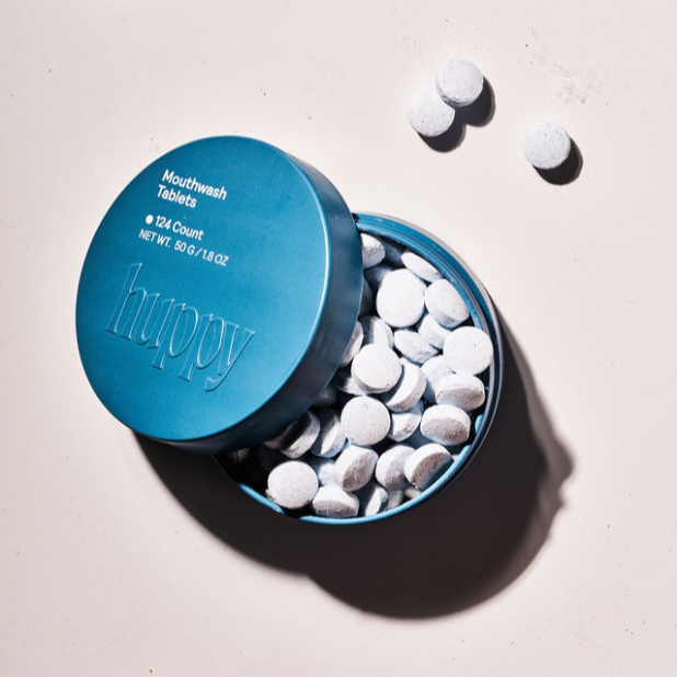 A round blue Huppy tin labeled Mouthwash Tablets features plastic-free packaging and contains white tablets, some scattered beside the open container on a light surface, emphasizing an eco-friendly oral care routine.