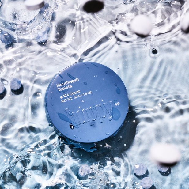 A round blue, plastic-free Huppy tin labeled Mouthwash Tablets floats in clear water, surrounded by scattered white and blue tablets, emphasizing a fresh approach to oral care.