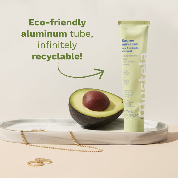 A tube of Mustela balm is displayed vertically next to a halved avocado on a tray with jewelry. The text beside the tube reads, "Eco-friendly aluminum tube, infinitely recyclable!" indicating its environmentally friendly packaging.
