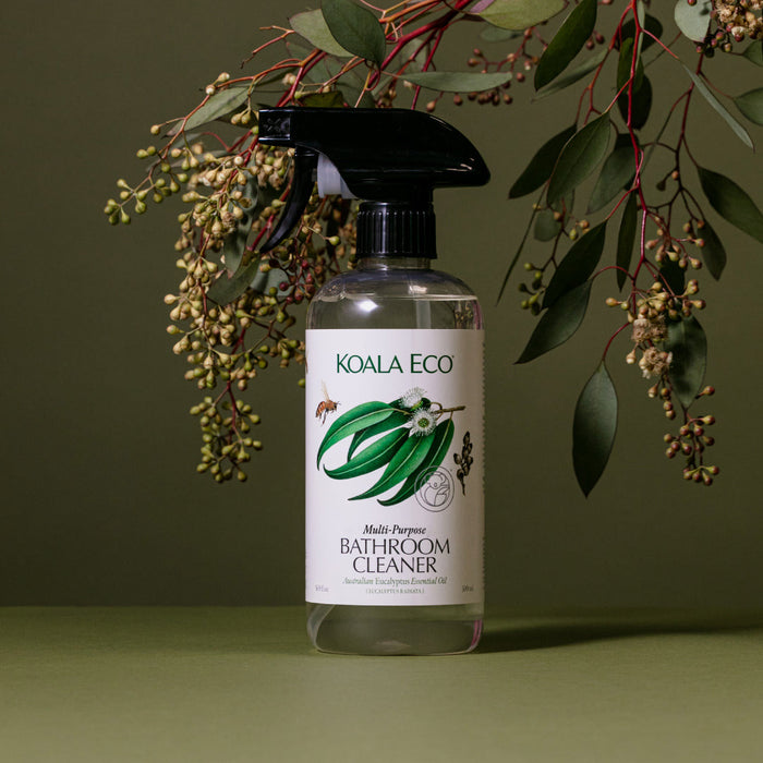 A spray bottle of Koala Eco Multi-Purpose Bathroom Cleaner rests on a green surface with eucalyptus branches in the background. The label highlights its effectiveness as a soap scum remover made with natural eucalyptus essential oil.