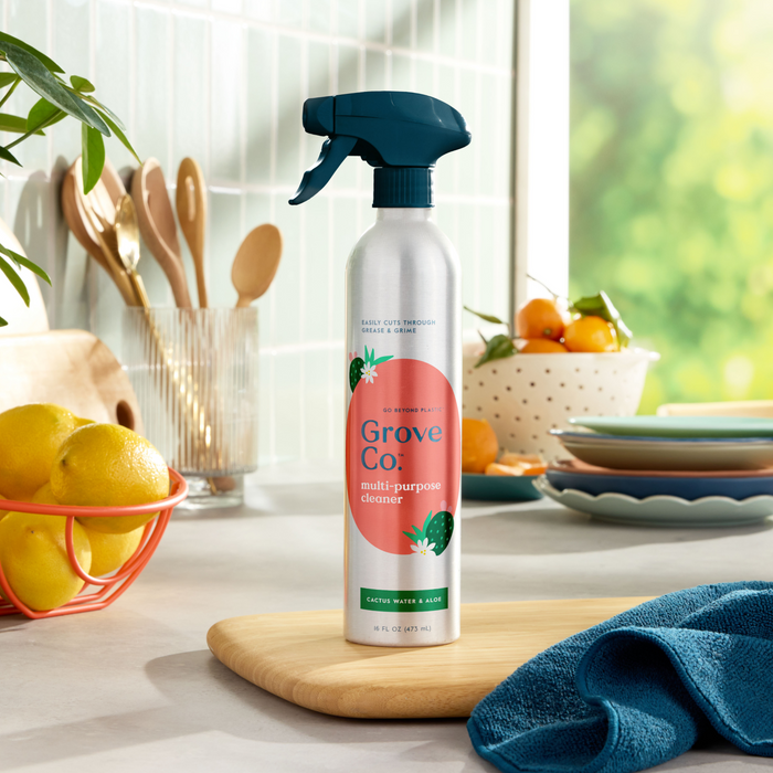 A kitchen counter displays a Grove Co. Multi-Purpose Cleaner (VIP Discount) bottle on a wooden board. Nearby, recyclable aluminum spray bottles shine among lemons, a blue cloth, colorful plates, wooden utensils, and a plant. Bright and airy with natural light, it’s ready to tackle household messes.