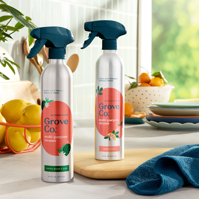 Two bottles of Grove Co.s Multi-Purpose Cleaner (VIP Discount) in recyclable aluminum spray bottles with a cactus water and aloe label sit on the kitchen counter. Nearby, a bowl of oranges and lemons complements stacked plates, while a blue cloth catches sunlight through the window.