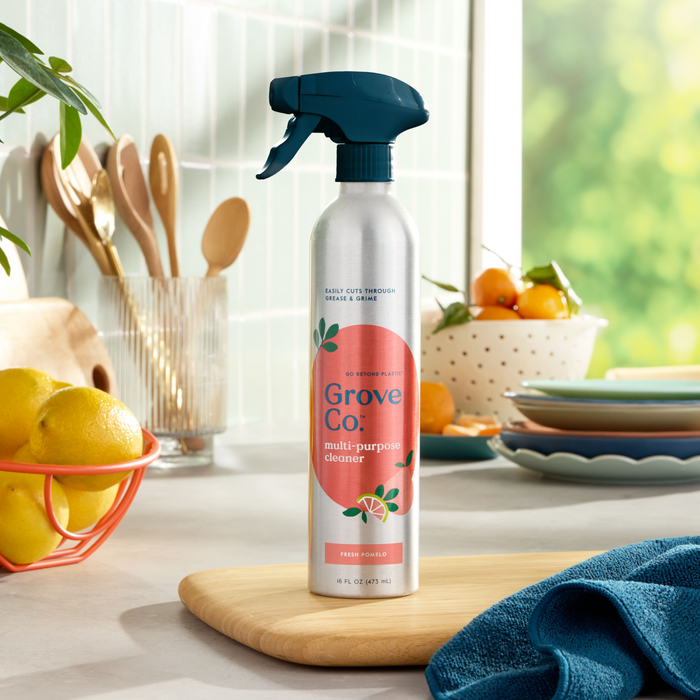 A Grove Co. Multi-Purpose Cleaner (VIP Discount) in a recyclable aluminum spray bottle is on the counter with a cutting board and cloth. Stacked plates, lemons, wooden utensils, and oranges bathe in sunlight through the window, ideal for tackling household messes.