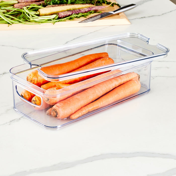 An iDesign Multi-Purpose Divided Bin - With Tray made with Recycled Plastic, filled with whole carrots, rests on a marble countertop with its lid partially open. In the background, a cutting board holds more carrots and a knife, hinting at meal prep in progress.