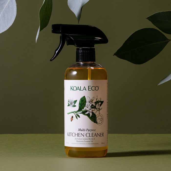 A bottle of Koala Eco Multi-Purpose Kitchen Cleaner with a black spray nozzle sits on a green surface, surrounded by green leaves and a green background. The label features botanical illustrations of Lemon Myrtle and Mandarin.