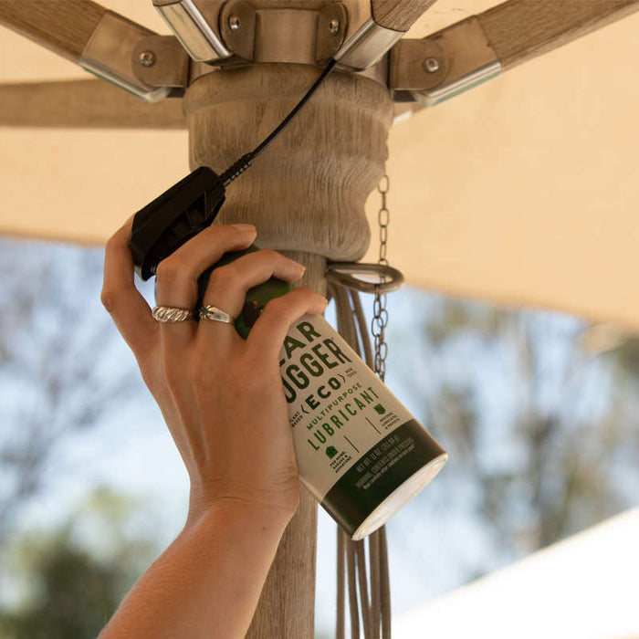 A hand uses Gear Huggers Multi-Purpose Lubricant spray on a patio umbrella mechanism. The sky and umbrella fabric are in the background, aiding smooth operation and rust protection.