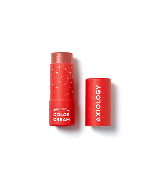 A red Axiology Multi-Stick Color Cream, cap off, reveals the product. The plastic-free packaging displays small white triangles and text. Its vegan and cruelty-free cap rests beside it on a spotless white surface.