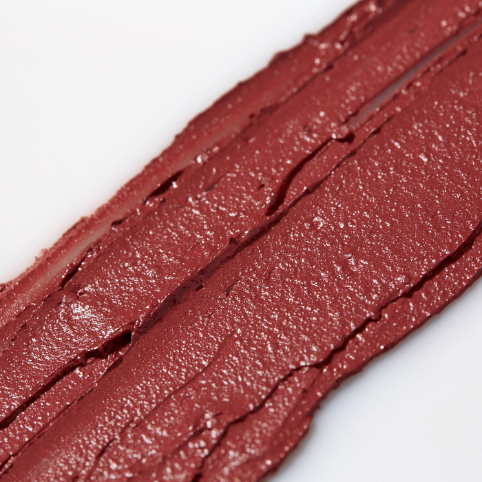 A close-up of Axiologys Multi-Stick Color Cream smeared on a white surface highlights its creamy texture, shiny finish, and visible grooves. This vegan, cruelty-free formula comes in plastic-free packaging, making it a bold and eco-friendly choice.