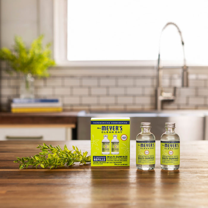 Two bottles of Mrs. Meyers Clean Day multi-surface cleaner with a lemon verbena scent sit on a wooden kitchen counter next to a green box of Mrs. Meyers Multi-Surface Concentrate Refills, while a potted plant and kitchen sink add charm to the cozy scene in the background.