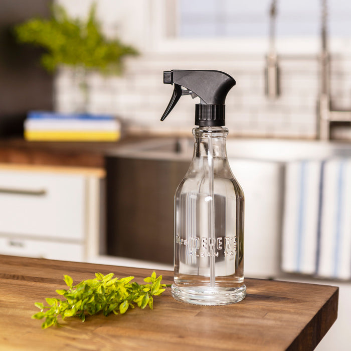 A Mrs. Meyers Multi-Surface Concentrate Refills bottle with a black trigger nozzle sits on a wooden kitchen counter, emitting garden-inspired scents. Fresh green herbs are in front, while a stainless steel sink, tiled backsplash, and blurred kitchen items fill the background.