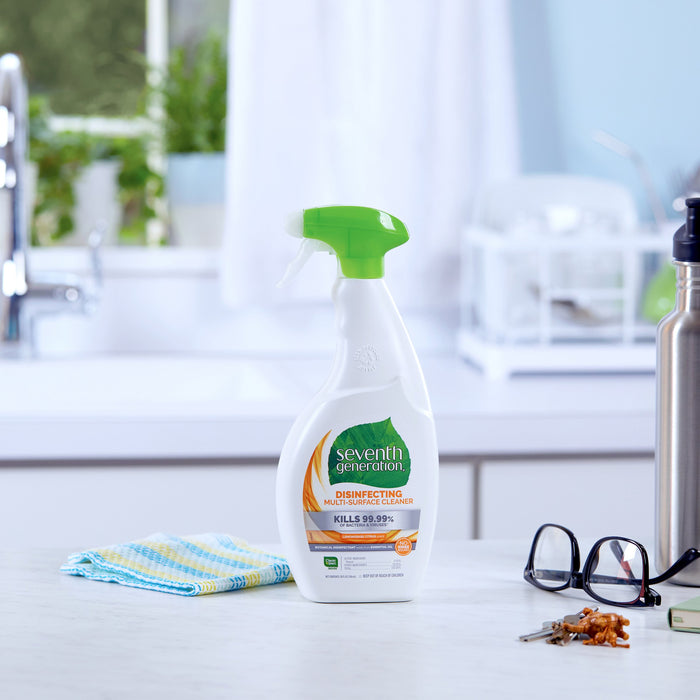 On the white countertop, a bottle of Seventh Generation Multi-Surface Disinfectant Cleaning Spray with a green nozzle exudes lemongrass citrus. Nearby rest glasses, a folded cloth, and a stainless steel water bottle. A small plant adds life to the scene.