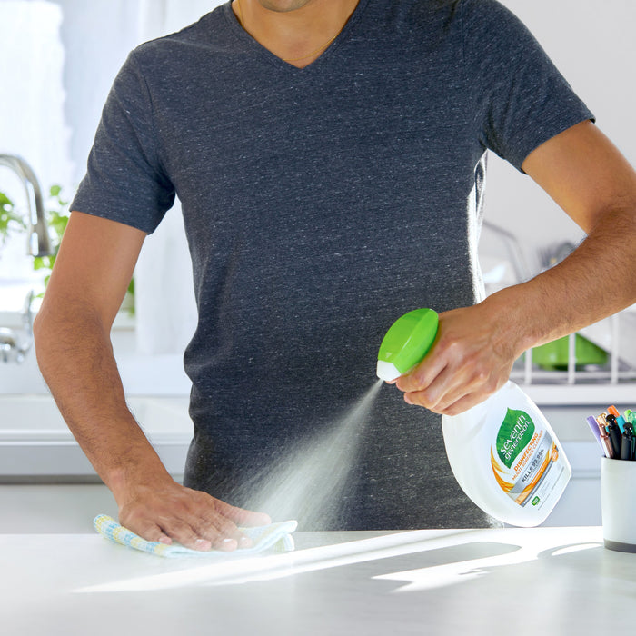 A person in a gray T-shirt uses a Seventh Generation Multi-Surface Disinfectant Cleaning Spray, with a green nozzle, to clean a brightly lit kitchen countertop. The room fills with a refreshing lemongrass citrus scent as they wipe the surface with a cloth; a sink and window are visible in the background.