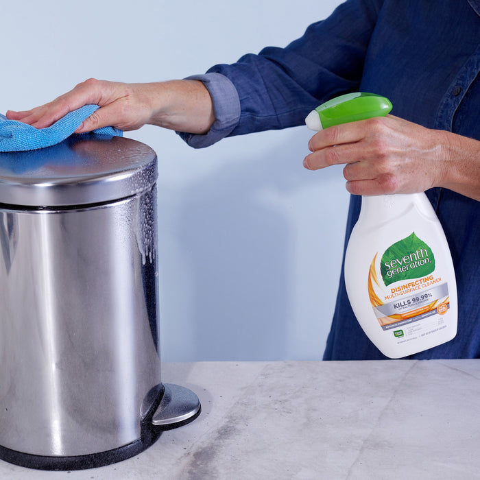 Wearing a denim shirt, someone cleans a stainless steel trash can using Seventh Generations Multi-Surface Disinfectant Cleaning Spray with Lemongrass Citrus scent and a blue cloth. The spray effectively kills 99.9% of germs.