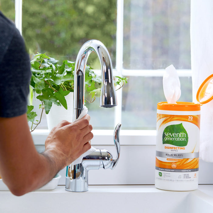 A person cleans a shiny kitchen faucet with Seventh Generation Multi-Surface Disinfecting Wipes, exuding a Lemongrass Citrus scent. Nearby, the container sits on the counter as a potted plant and bright window form a serene backdrop.