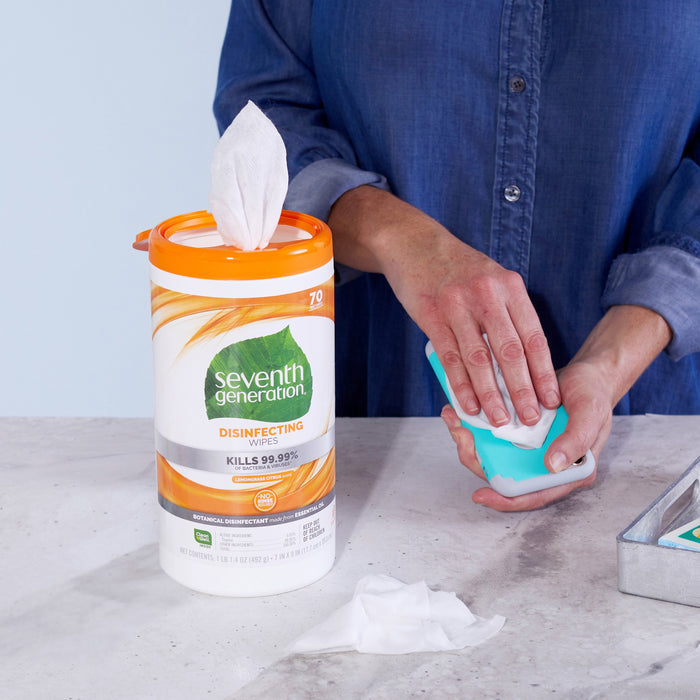 A person in a blue shirt uses Seventh Generations Multi-Surface Disinfecting Wipes from a container promising to kill 99.9% of germs with Lemongrass Citrus scent, cleaning a small teal object. Disposable wipes sit on the countertop, ready for multi-surface use.